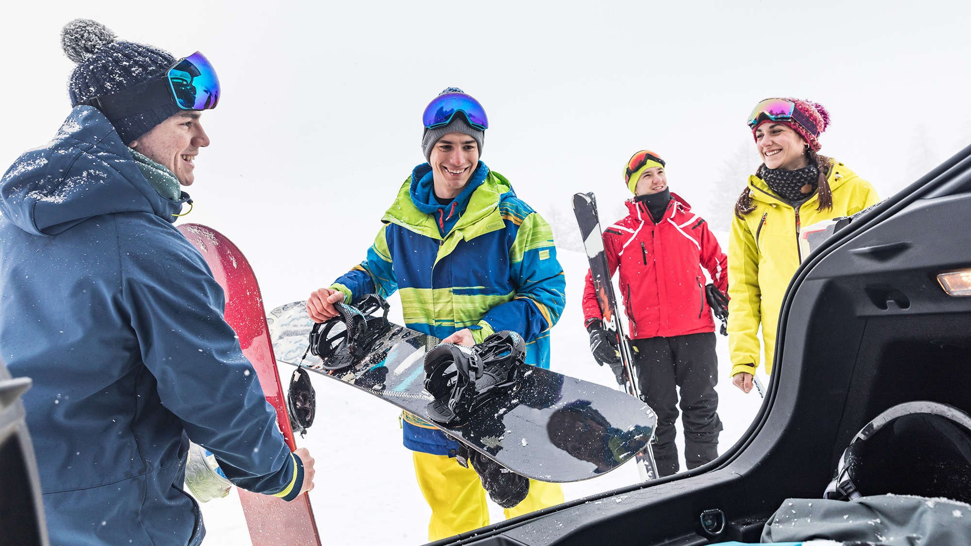 2 men and 2 female friends dressed in ski/winter coasts and clothing, loading ski gear and snowboards into the boots of a car